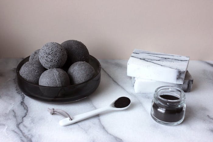 black bath bomb ideas, small jar and spoon, both filled with black powder, on a marble surface, with black dish, containing several dark gray bath bombs
