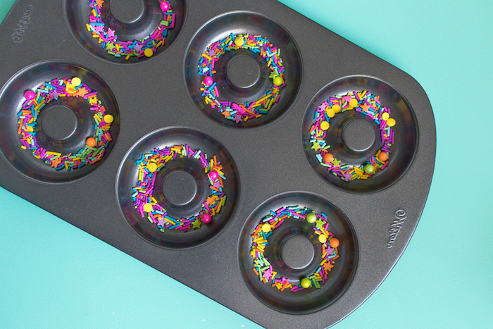 teflon dish in black, with donut-shaped moulds, containing multicolored sprinkles, bath bombs recipe ideas, pale blue background