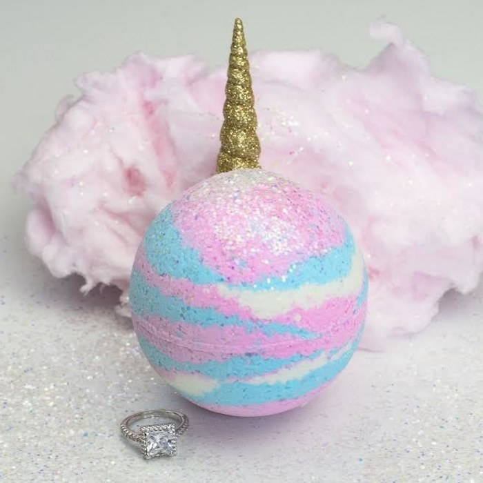 cotton candy in sparkly pale pink, behind a bath bomb in white, baby pink and blue, with silver glitter, and a unicorn horn, covered in golden glitter, a silver ring with large stone nearby