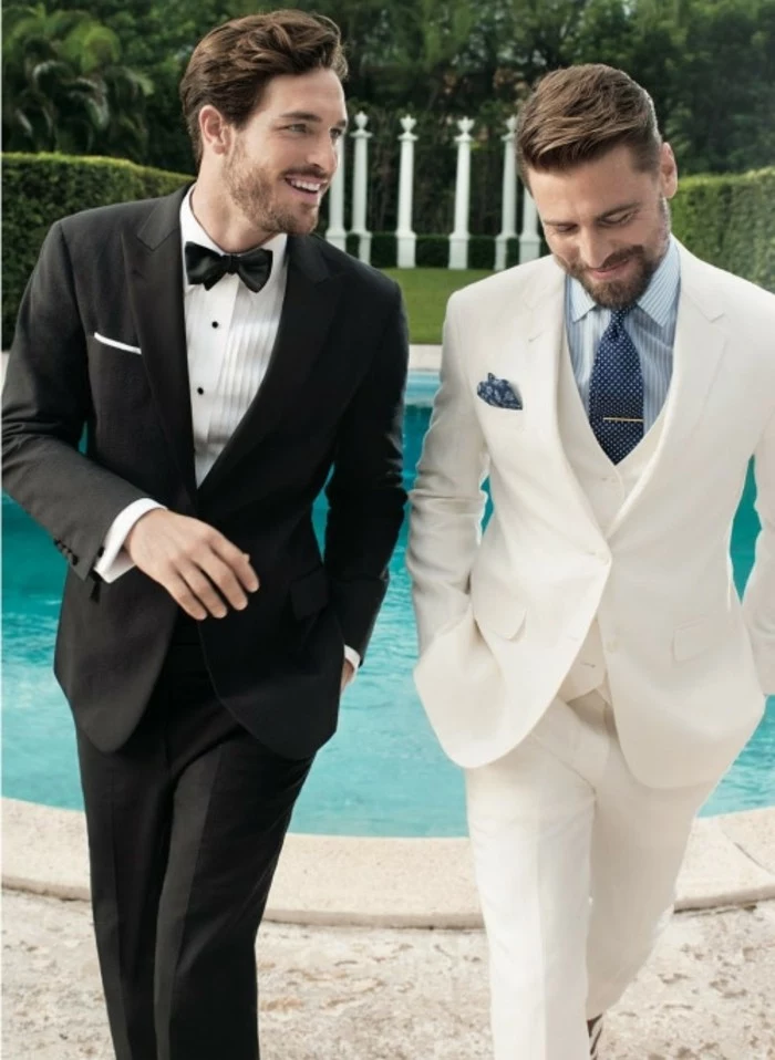 talking and smiling men, one wearing a black tuxedo, with white shirt and black bowtie, how to dress for a wedding male, the other in a white three piece suit, with light blue shirt, and dark blue tie