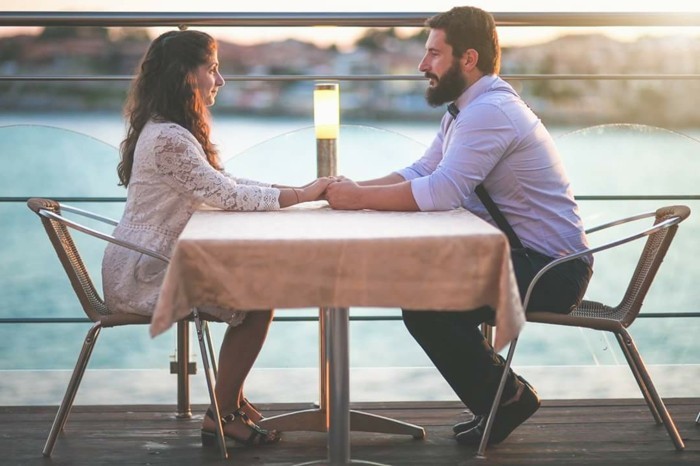 table near the sea, with a couple holding hands, her dressed in a white lace dress, him wearing a white shirt, dark trousers and a tie, how to dress for a wedding male