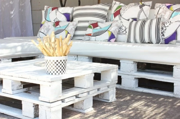 matching white pallet table, and corner sofa, pallet patio furniture, white cushions in different sizes, decorated with stripes, and colorful patterns