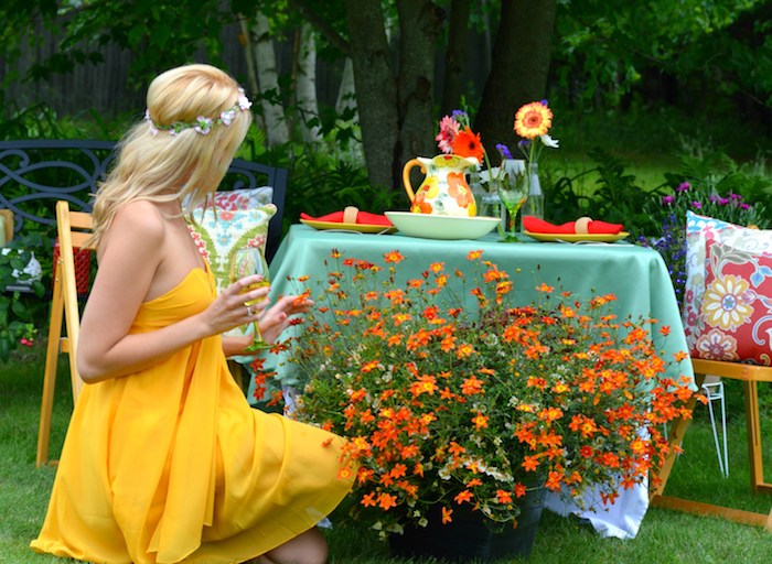 platinum blonde woman, with small flower crown, dressed in floaty, vibrant yellow strapless dress, admiring orange potted plants in a garden, while holding a glass of wine