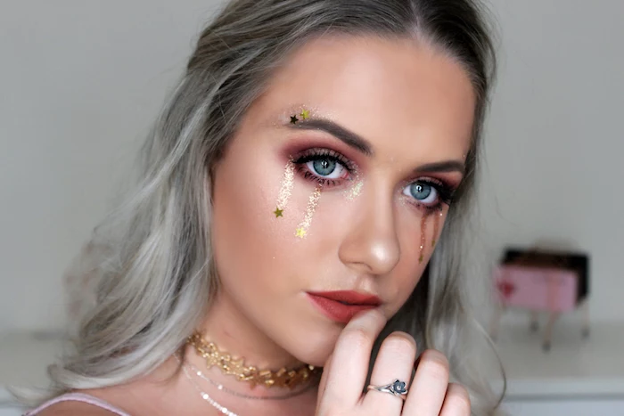 golden tears made from body glitter, with little star stickers, decorating the face of a woman, with maroon eye makeup, red lipstick and grey ombre hair