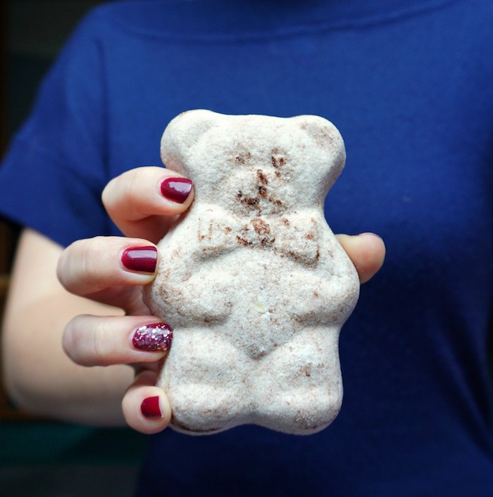 hand with dark red nail polish, holding a large beige bath bomb, shaped like a teddy-bear, funny bath balls ideas