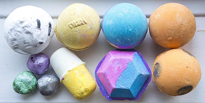 pill and gem-shaped bath bombs, near several round bath balls, in different sizes and colors, white and yellow, blue and orange, green and violet