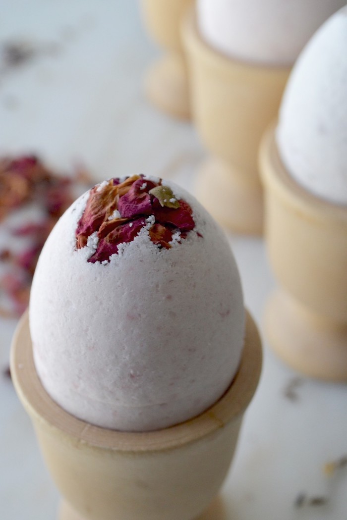 close up of a pale pink bath bomb, shaped like an easter egg, and decorated with dried dark pink rose petals, bath bomb recipe, wooden egg dish