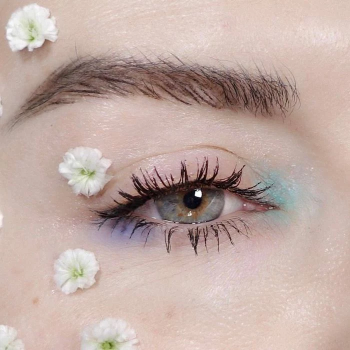 daubs of teal and pale violet eyeshadow, on the corners of a blue eye, with black mascara, cute makeup looks, several small white and green blossoms surround the eye