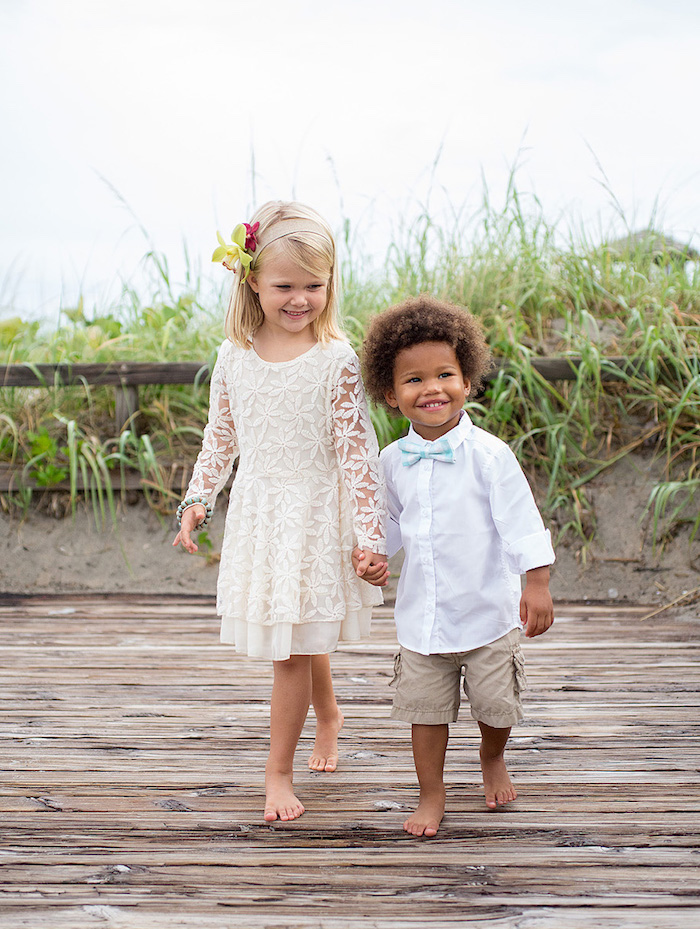 children holding hands and smiling, little boy in white shirt, khaki shorts and pale blue bowtie, little girl in a white embroidered dress, with flowers in her hair