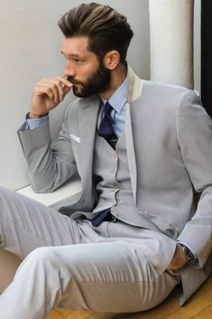 bearded brunette man, in light gray three piece suit, with light blue shirt, and a dark blue tie, sitting with a hand in his pocket