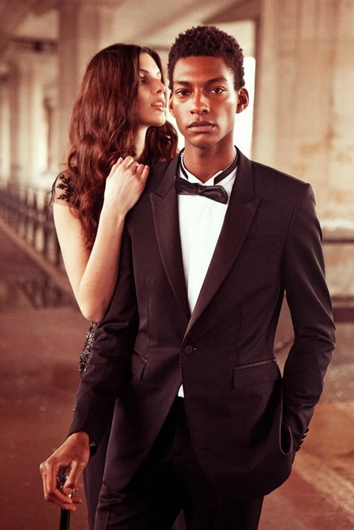 mens wedding guest attire, young black man, wearing a black tuxedo, with a white shirt, and a black bowtie, a cane in his hand