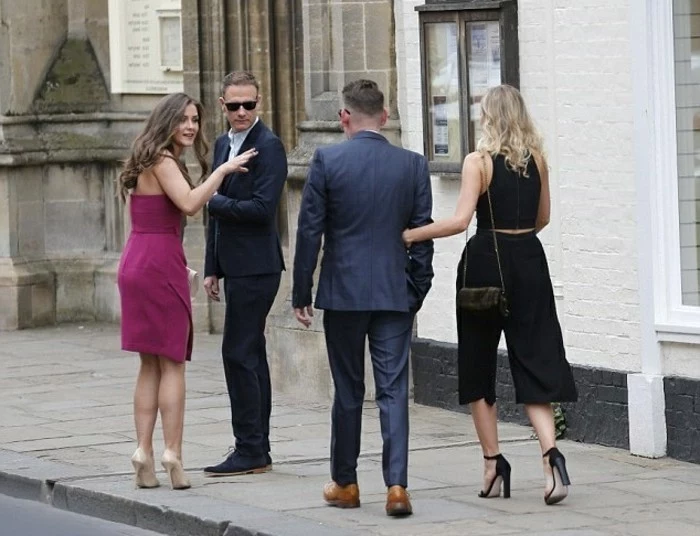 four people on a street, brunette woman in purple dress, blonde woman wearing black culottes and a top, and two men in dark suits, mens summer wedding attire, sunglasses and other accessories