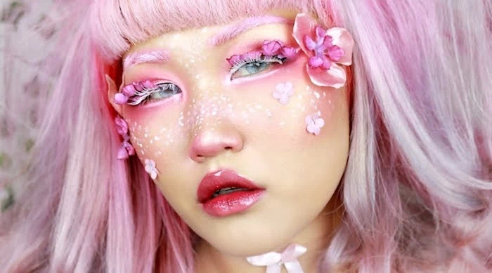 doll-like artistic make up, glossy pinkish-red lipstick, fake eyelashes with white mascara, pink and white face paint, faux flowers stuck on the sides of the face, pastel pink hair with short cropped bangs