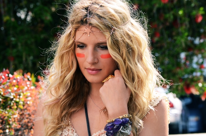 stripes of red paint, decorating the cheeks of a blond woman, with curly hair and dark roots, wearing a rose gold headdress, with a star shape