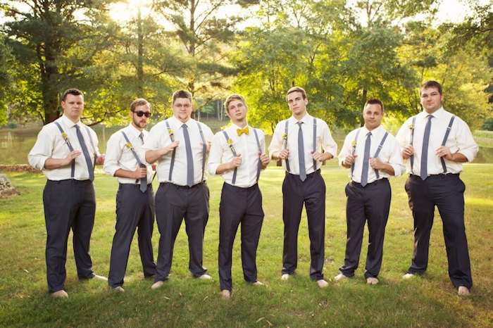 seven barefoot men, wearing identical outfits, black trousers and white shirts, with suspenders and grey neckties, one man is wearing a yellow bowtie, dressy casual men, standing in a garden with trees