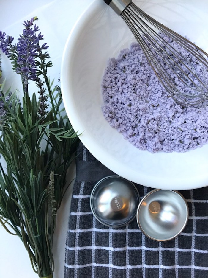 homemade bath bombs, coarse lumpy violet powder, inside a white ceramic bowl, with a whisking tool, metal moulding dish and several violet flowers