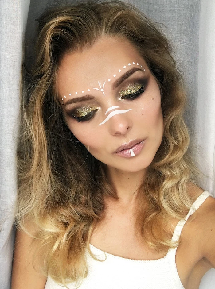 slim woman with wavy brunette hair, and blonde balayage, wearing brown and sparkly golden eyeshadow, makeup looks, glossy nude beige lipstick, and white face paint