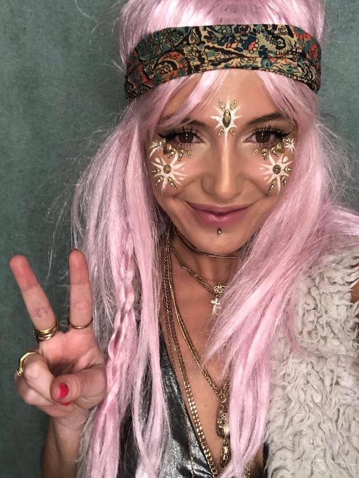 hippie costume with pastel pink long wig, decorated with small messy braid, and multicolored patterned headband, worn by smiling woman, with gold and white facepaint