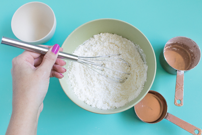 pink nail polish, on woman's hand, holding a whisking utensil, and mixing white powder, in a pale green plastic bowl, making bath bombs, white bowl and measuring spoons nearby