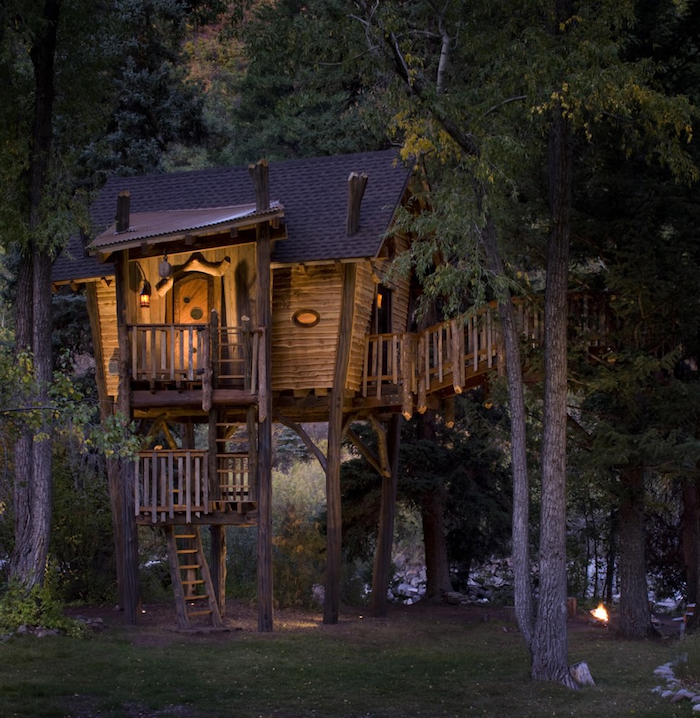 asymmetrical wooden house, built above the ground, and supported by several trees, lit from within, photo taken at night