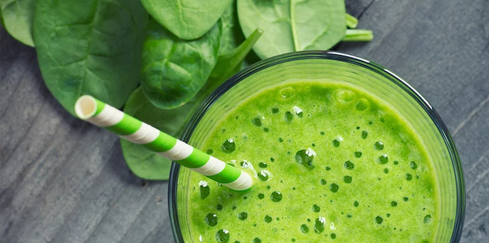foamy light green smoothie, inside a tall clear glass, with a white and green striped paper straw, healthy breakfast ideas, next to spinach leaves 