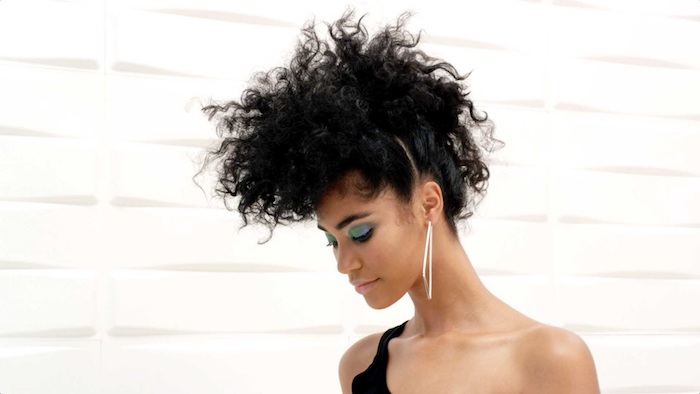 mohawk-like black hair, styled from afro curls, and worn by young woman, with light blue eye make up, black one shoulder top, and large triangular earnings, hairstyles for curly hair 