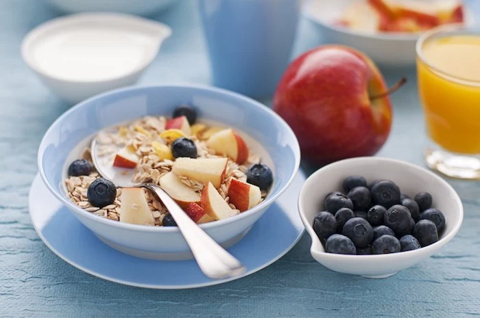 whole apple and a glass of orange juice, near a bowl containing a spoon and rolled oats, apple chunks and blueberries