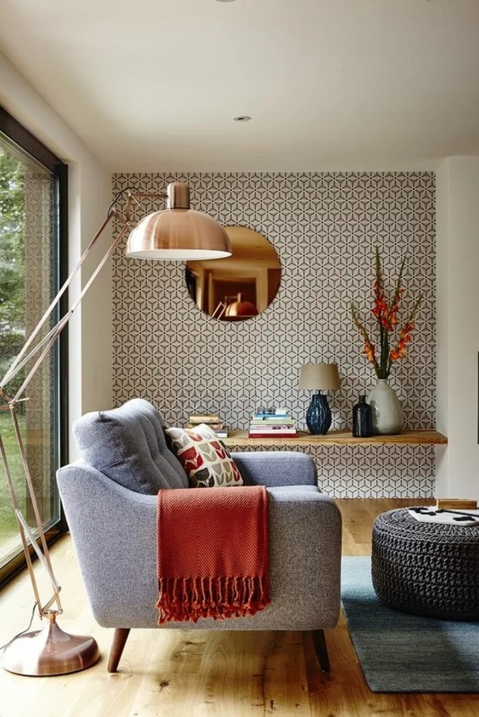copper-colored lamp, standing over an armchair in grey, with a patterned cushion and a red throw, on a beige wooden floor, near a faded blue rug, and a coffee table
