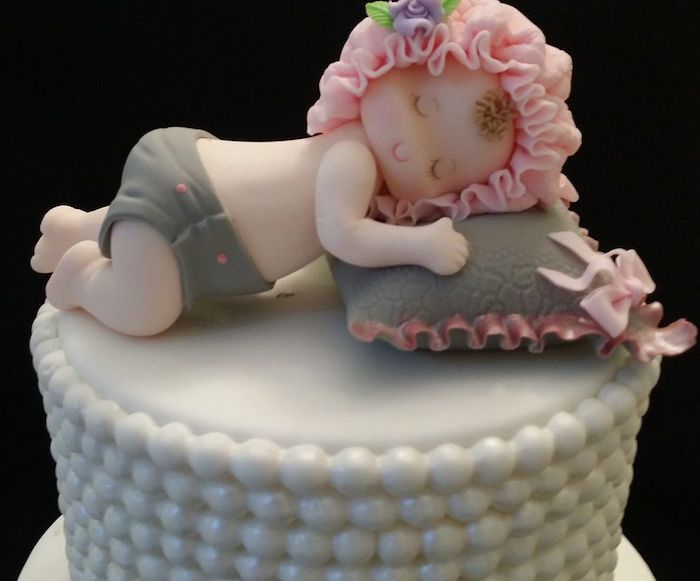 smiling sleeping baby figurine, resting a grey and pink pillow, baby shower cake toppers girl, with grey diaper, and a pale pink cap, with a tiny purple flower, made from fondant