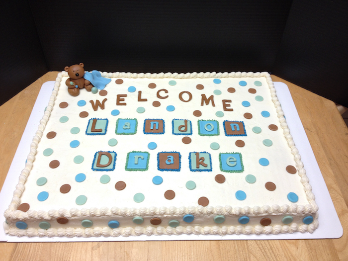 teddy bear figurine holding a blanket, made from brown and blue fondant, decorating a large cream-colored cake with blue, brown and turquoise details, baby shower sheet cakes 