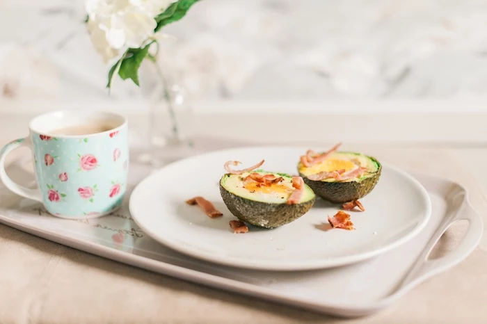 avocado cut in two halves, with egg and bacon flakes, on a white round plate, breakfast menu ideas, cup of milk tea or coffee next to it