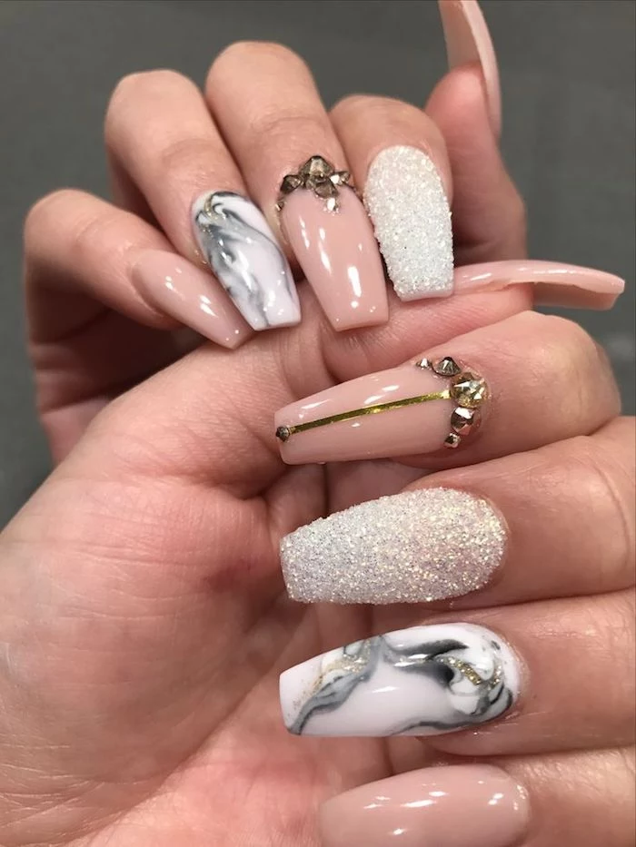 rhinestones in gold and white textured glitter, decorating the coffin shaped nails, of two hands, pale nude pink nail polish, and white nail polish decorated with grey, marble-like patterns
