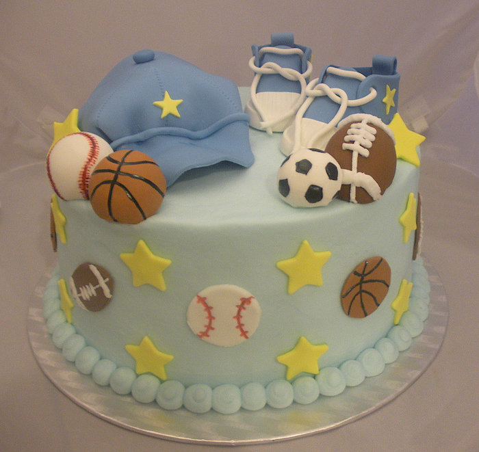football and soccer, basketball and baseball balls, made from fondant, and placed near a pale blue baseball cap, and little pale blue sneakers, on top of a light turquoise cake, decorated with balls, and yellow stars