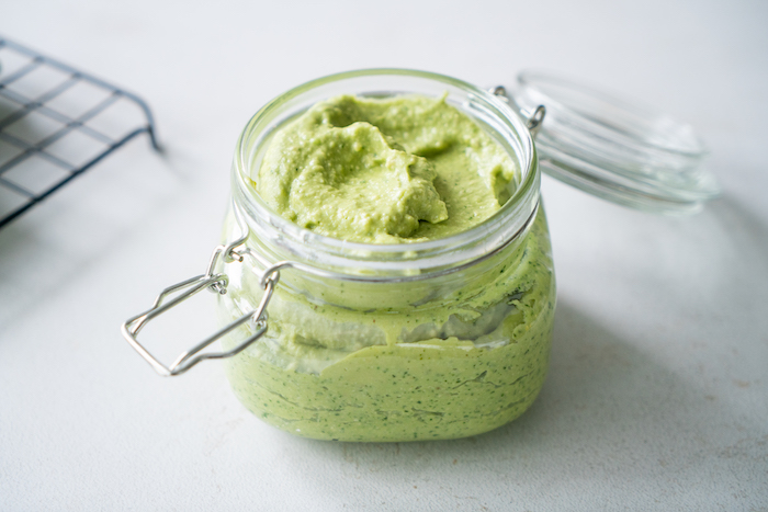 basil pesto with avocado, medium glass jar with lid, filled with pesto, placed on white surface