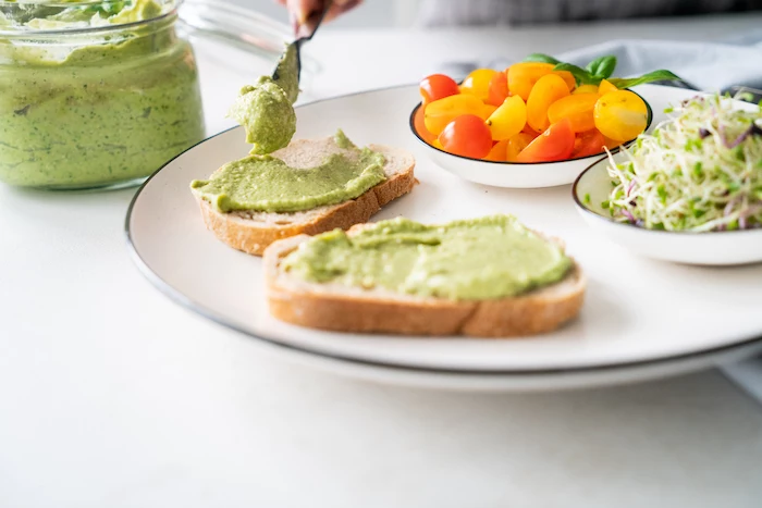 cherry tomatoes in a bowl, basil pesto with avocado, spread on two bread slices, placed on white plate