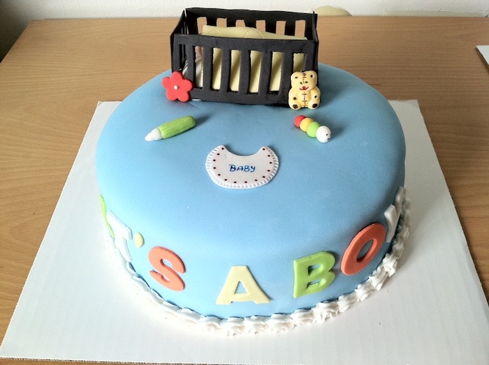 crib in dark brown, made from fondant, with a yellow blanket, a teddy bear and other baby items, on a cake with smooth blue frosting, baby shower cakes for boys, decorated with the words it's a boy