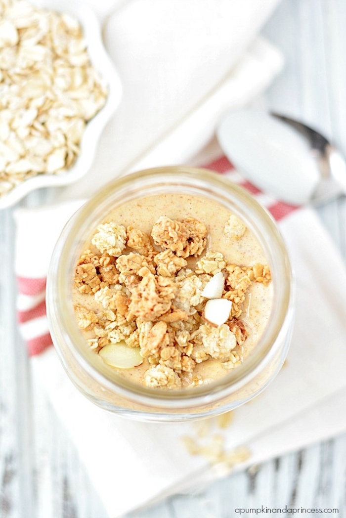 granola and nuts, strewn on top of a pale orange porridge, inside a glass jar, simple breakfast ideas, small bowl with rolled oats, and metal spoon nearby