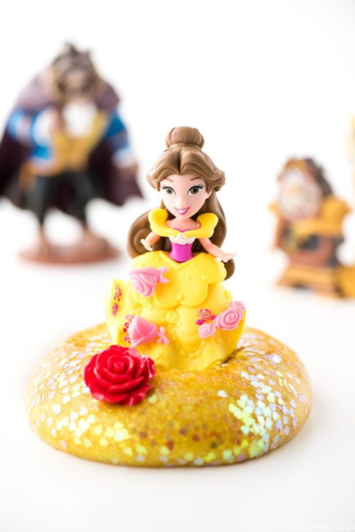 beauty and the beast-themed slime without borax, plastic character figurine, in yellow and pink dress, with brown hair, standing on a pile of yellow goo, covered with iridescent glitter flakes