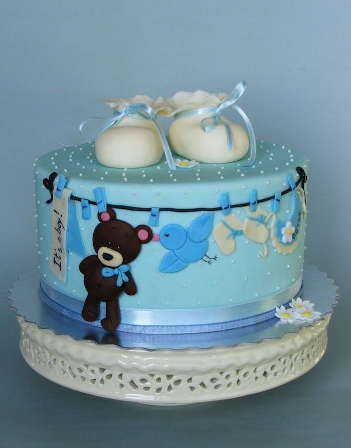 clothes line with a brown teddy bear, little white shoes, a baby's bib, and other items, decorating a pale blue cake, topped with a pair of white baby booties, with pale blue ribbons