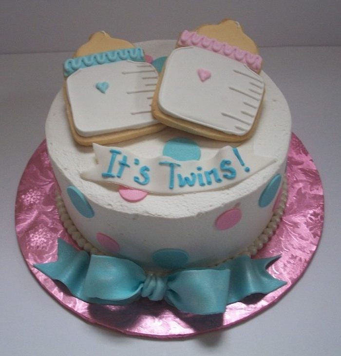 it's twins written in blue frosting, on a white fondant ribbon, decorating a white cake, twin baby shower cakes, topped with two cookies, shaped like baby bottles