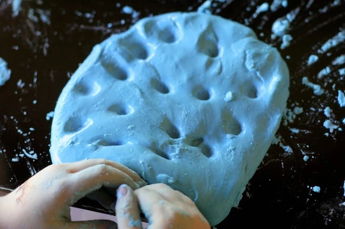 holes poked into a glossy, smooth pile of light blue slime, placed on a dark surface, two tiny child's hands, covered in blue slime nearby