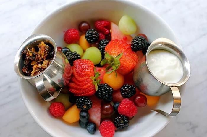 healthy low calorie breakfast, fruit salad with strawberries, blackberries and blueberries, watermelon slices and grapes, inside a white bowl, with small dishes of yoghurt and granola 