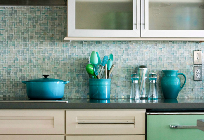 turquoise and pale blue, glass mosaic tile backsplash, in a kitchen with cream, and pale green cabinets, and a dark grey counter top