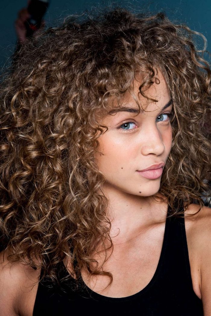 messy and voluminous, light brunette curly hair, with darker roots, highlights and curly bangs, worn by girl in black tank top, haircuts for curly hair, blue eyes and discrete make up