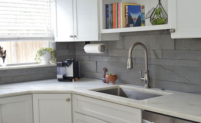 succulent in a gem-like container, and several books, on a white kitchen shelf, grey stacked stone backsplash, white cabinets and a marble counter top