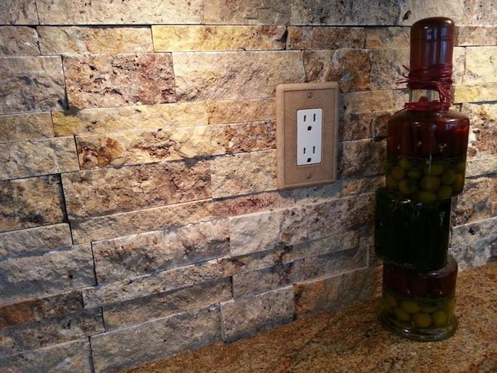 olives marinated in a bottle, on a beige spotted, stone-like counter top, near a stacked stone backsplash, in orange and beige, with a power outlet