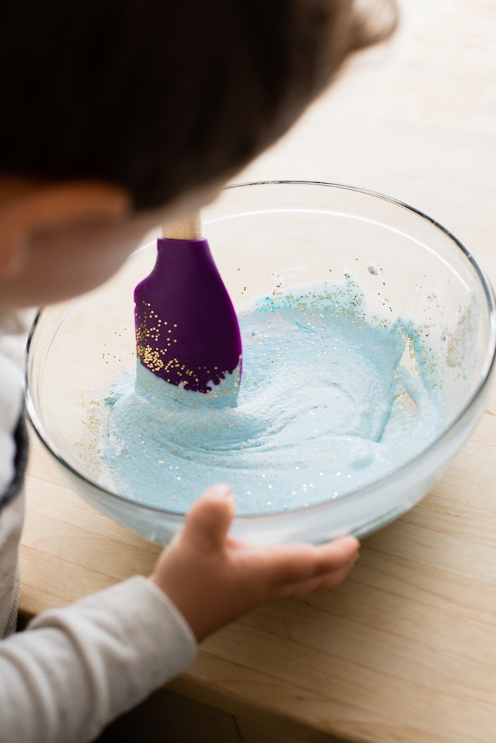 toddler holding a clear glass bowl, half-filled with a light blue, gooey mixture and glitter, fluffy slime recipe, mixing ingredients with a spatula