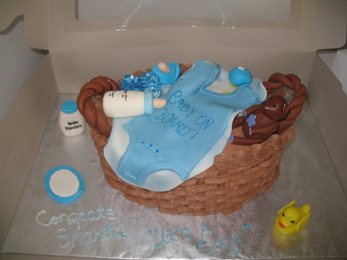 basket-shaped cake, made from brown fondant, and filled with baby items, teddy bear and bottle, dummy and blue onesie, all made from colored fondant, onesie cake with toys