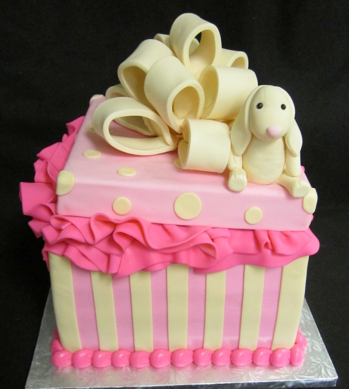 gift-shaped cake in pink, decorated with pale yellow fondant stripes, and polka dots, baby shower cakes for girls, with a large pale yellow bow, and a bunny toy fondant figurine