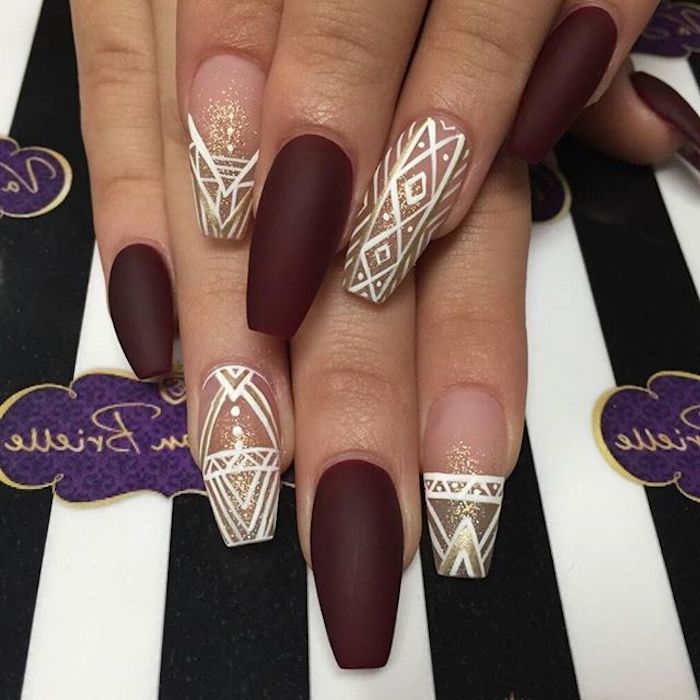 wine red matte nail polish, and clear glossy nail polish, on coffin acrylic nails, decorated with gold glitter, and white motifs, on two hands, one resting on top of the other